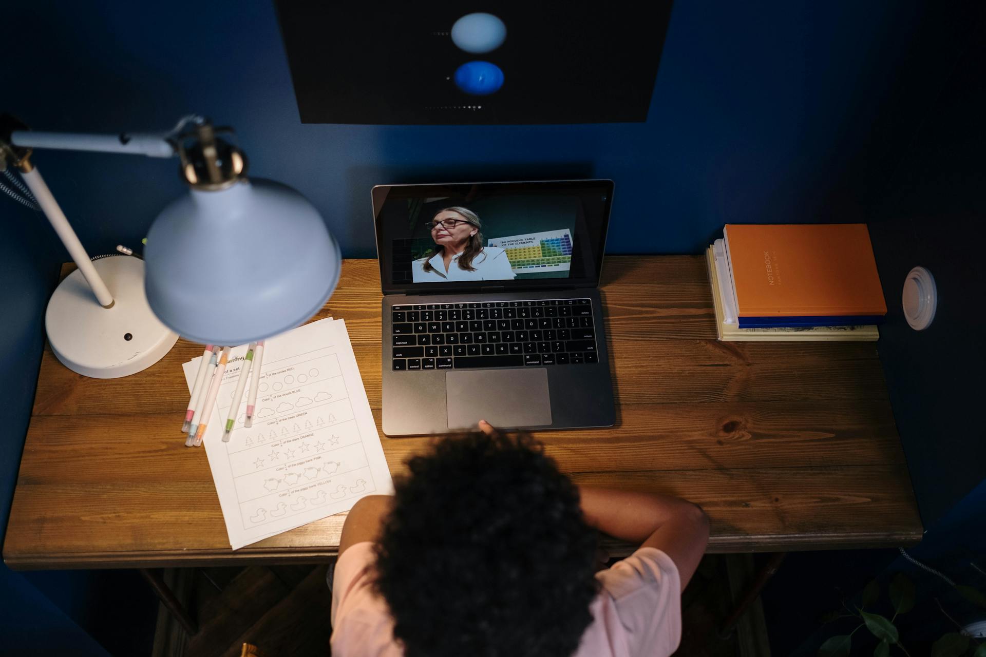 A student attends a virtual class, studying at a wooden desk filled with educational materials.