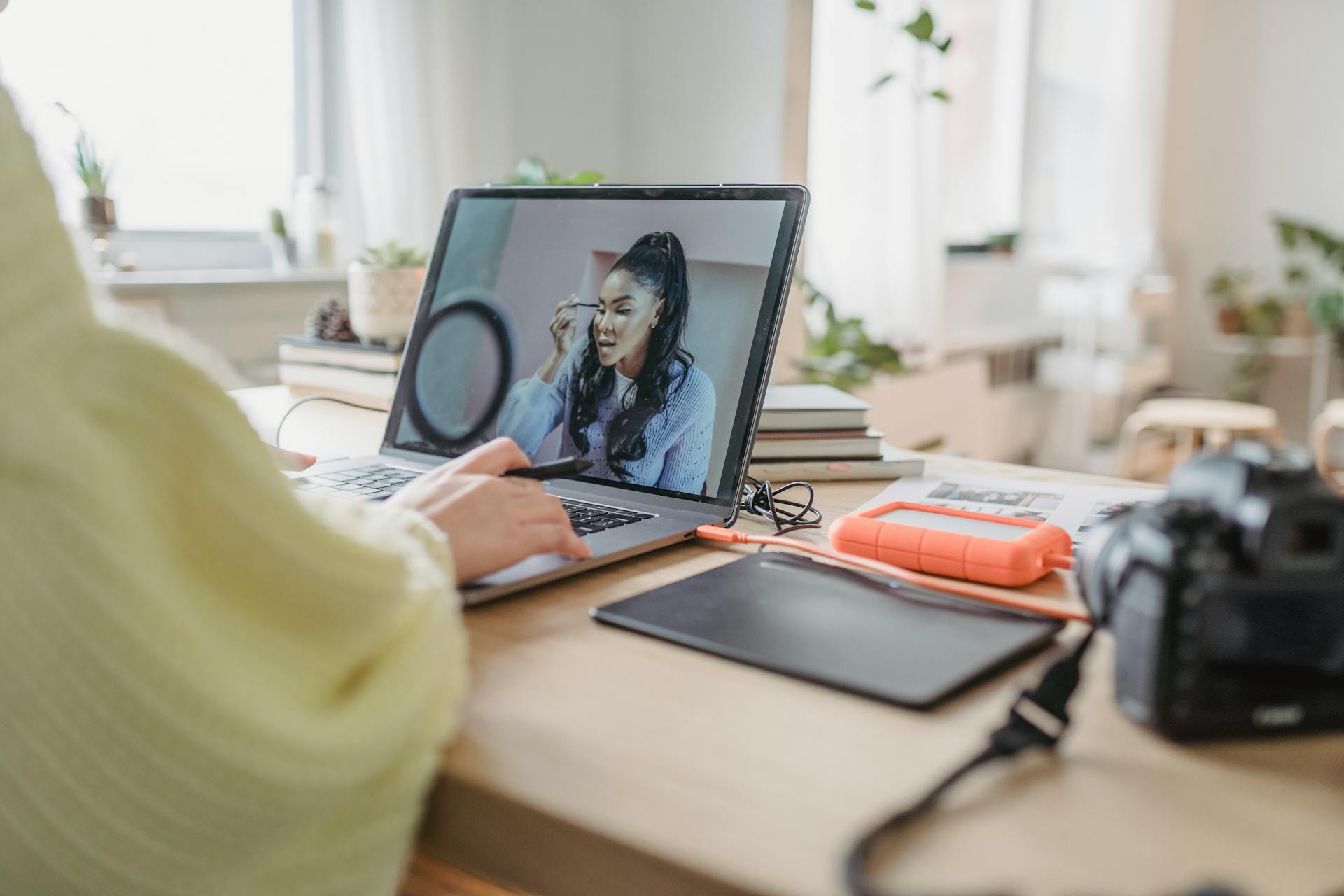 Crop unrecognizable female photographer editing photo while sitting at table with photo camera and graphic tablet during work in spacious room with flowerpots