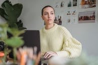 Serious female photographer with short hair looking away while browsing netbook at table with photo camera during work in room with green plants and printed photos