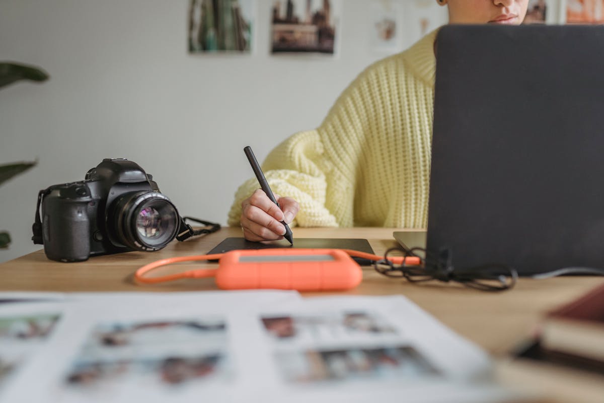 Crop anonymous woman using graphic tablet while editing photos on netbook at table with photo camera and blurred printed photos