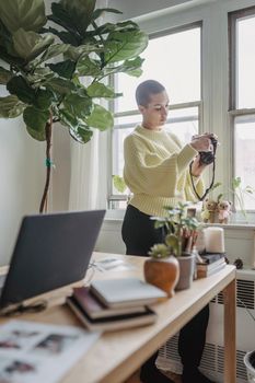 Side view of concentrated female photographer with short hair standing with professional photo camera in hands near table with modern netbook and flowerpots