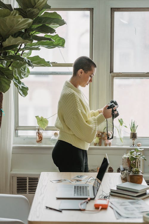 Side view of concentrated female photographer with short hair looking at screen of photo camera while standing between window and table with laptop and graphic tablet