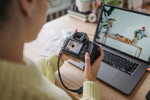 Free Unrecognizable crop female photographer looking at screen of photo camera while sitting at wooden table with netbook during work in light room Stock Photo