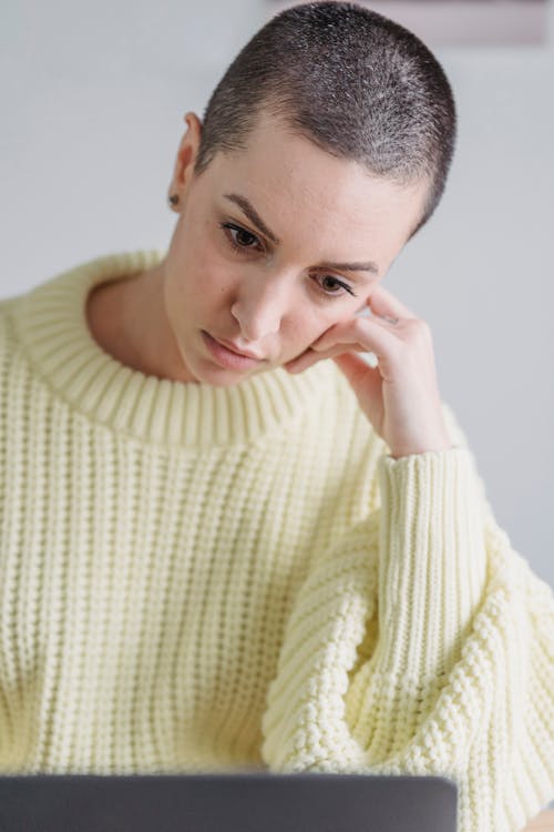 Free Serious female with short hair wearing warm yellow sweater browsing netbook while leaning on hand and sitting in light room Stock Photo