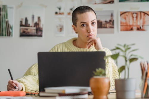 Serious female with short hair looking at camera while drawing with stylus at table with netbook and small potted plants