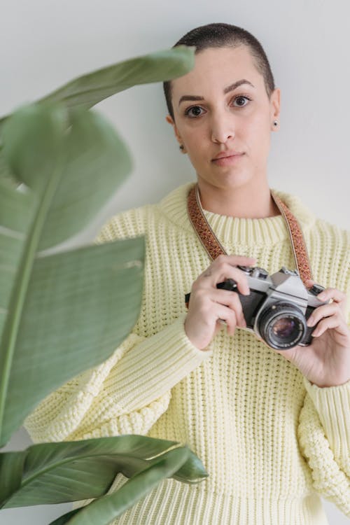 Confident female photographer with photo camera in hands looking at camera while standing on white background near green leaves of plant