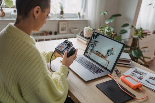 Side view of crop concentrated female photographer looking at screen of photo camera while sitting at table with netbook and graphic tablet