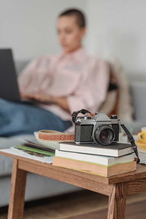 Free Modern photo camera placed on stacked books on coffee table near focused female working on netbook and lying on couch Stock Photo