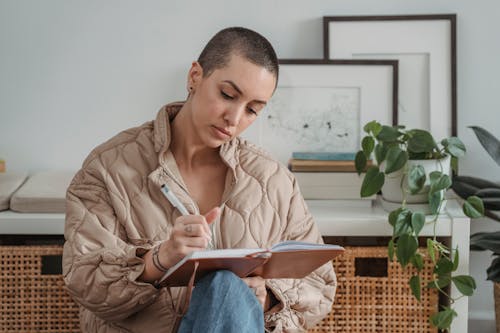 Pensive young female with short hairstyle writing in notebook while sitting in light living room