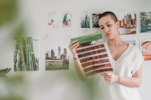 Thoughtful female photographer in white shirt looking at photo films in hands while standing near wall in light room