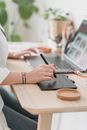 Crop anonymous female browsing netbook and working on graphic tablet with stylus while sitting at desk