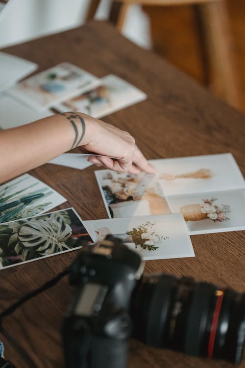 Crop unrecognizable photographer pointing at printed photos scattered on wooden table near professional photo camera
