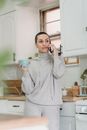 Content young female in domestic clothes having conversation via mobile phone and enjoying delicious coffee while standing in modern kitchen