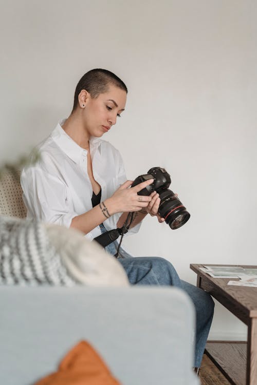 Pensive female photographer in casual wear setting professional photo camera and sitting on comfy couch in living room