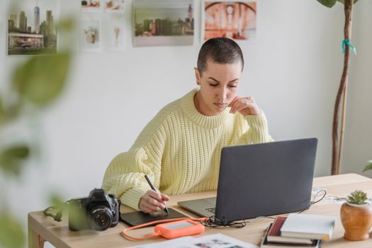Focused young female in casual sweater working on modern netbook and drawing on graphic tablet while sitting at desk