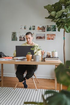 Full body of female freelancer in casual clothes sitting at table in light cozy workspace and working on laptop