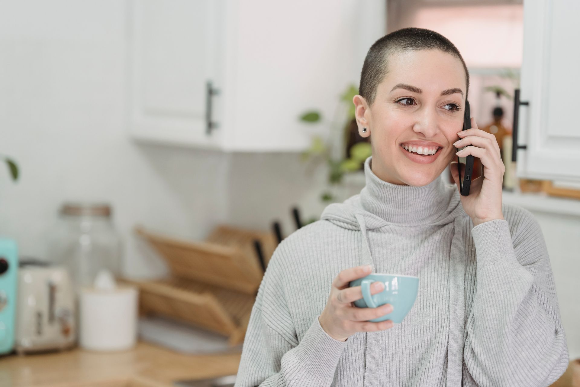 Female with short hair smiling and looking away while having smartphone conversation on blurred background of kitchen at home
