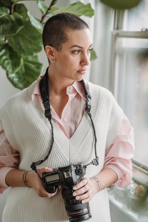 Thoughtful short haired woman standing with professional photo camera near window and looking away