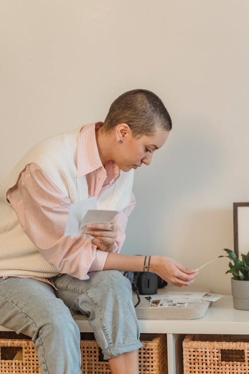 Young woman with shaved head sitting on cabinet with professional photo camera looking at printed pictures at home