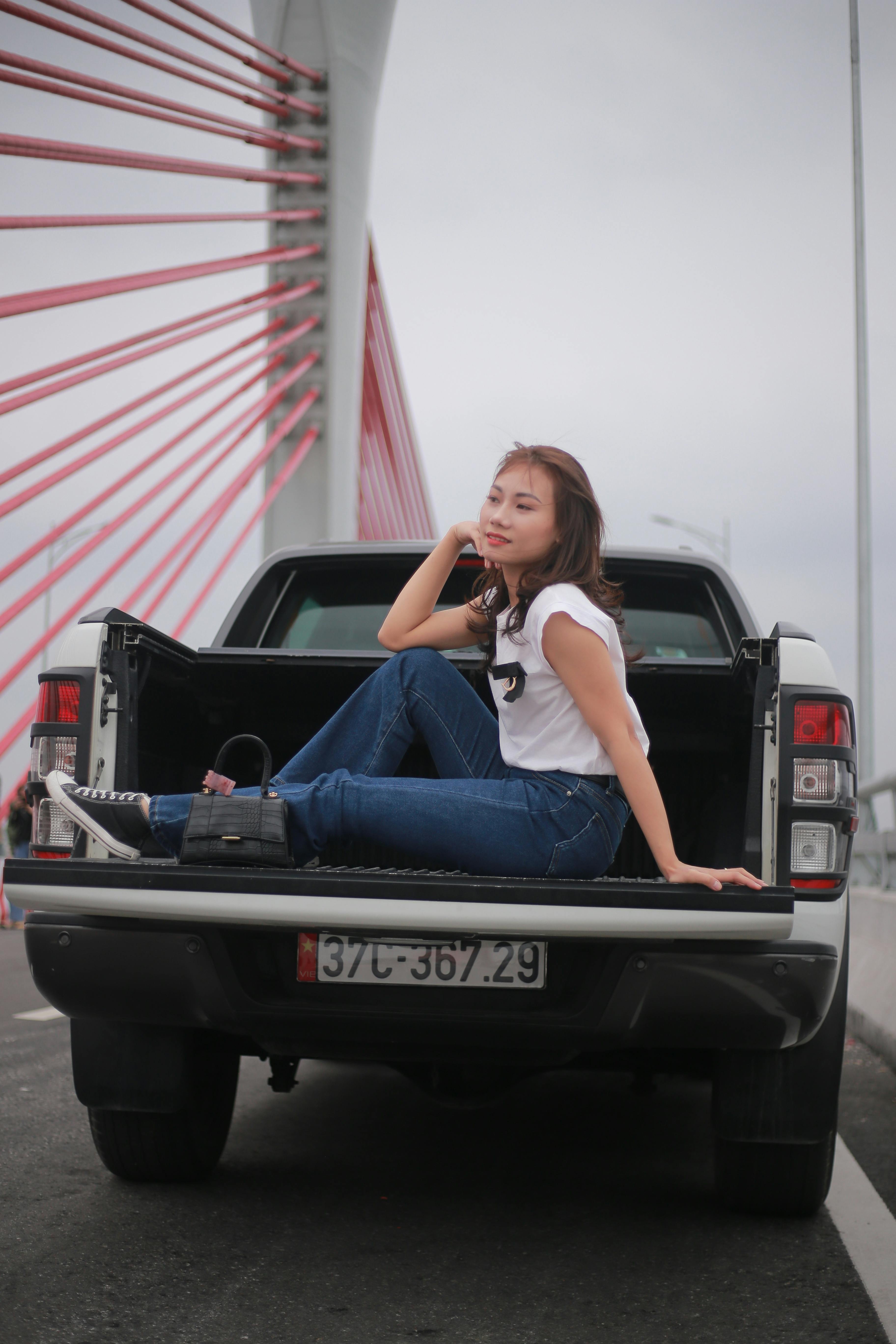 Asian woman in casual wear posing on a pickup truck parked on a modern bridge, full shot.