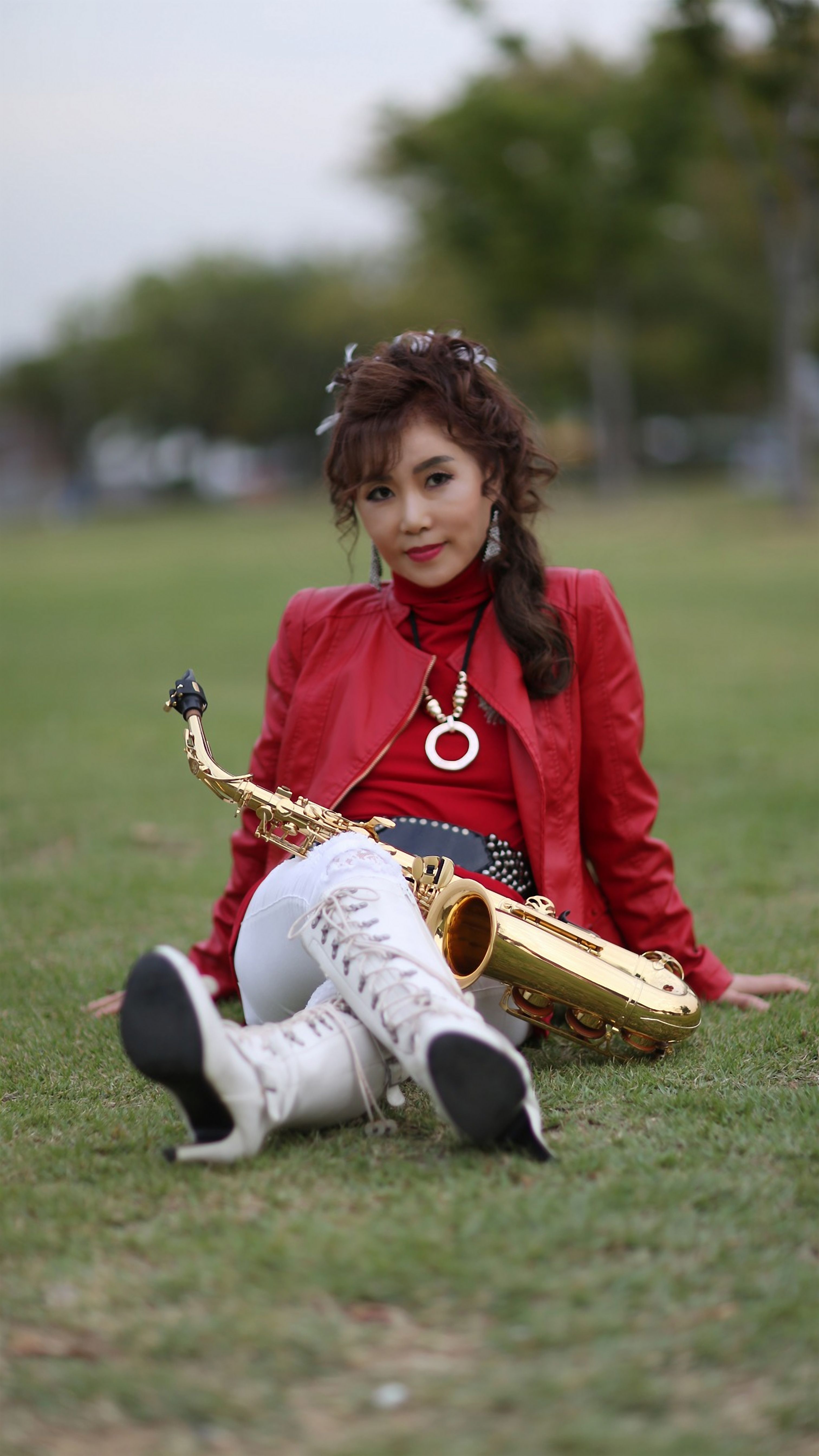 Woman in red jacket with saxophone sitting on grass in a park setting.