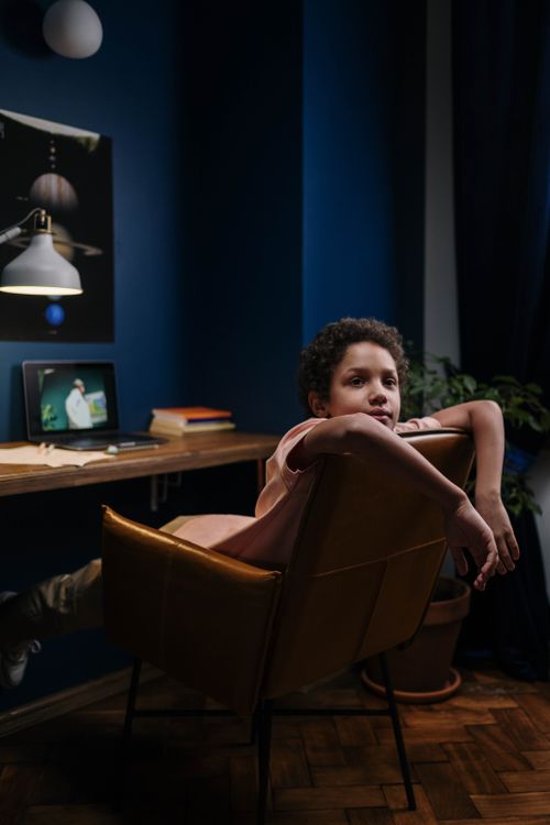 Free A child leans back in an armchair while attending an online class on a laptop in a cozy home setting. Stock Photo