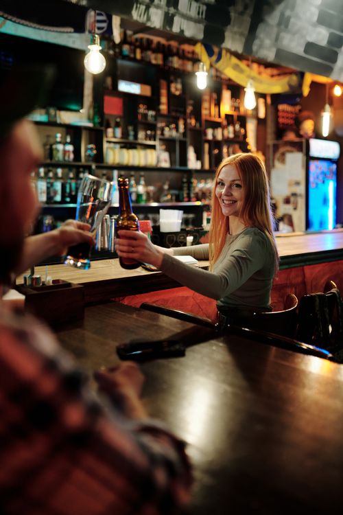 Free A cheerful young woman raises a beer bottle in a lively bar, embodying social enjoyment and night out vibes. Stock Photo