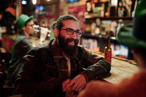 Free Bearded man in glasses chatting at a rustic bar with friends. Warm indoor ambiance. Stock Photo