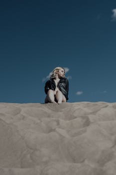 Woman in a leather jacket poses against a blue sky on a sandy dune.