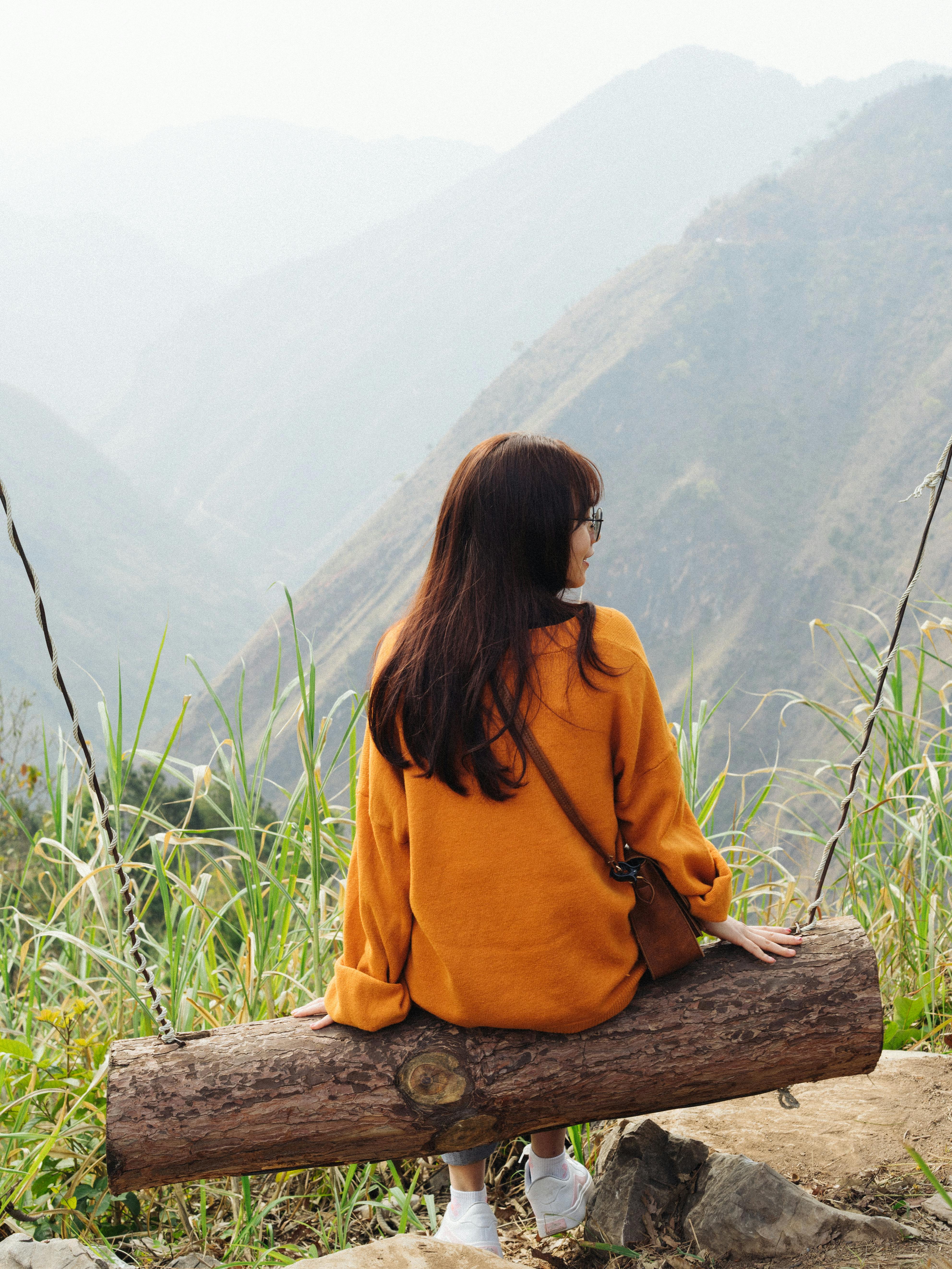 A woman in an orange sweater enjoys a scenic mountain view sitting on a log swing.