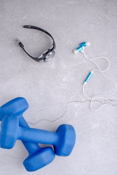 Flat lay of blue dumbbells, earphones, and a watch on a concrete surface.