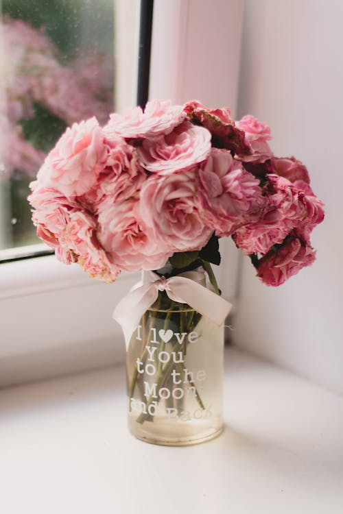 Bouquet of fresh rose arranged in transparent glass vase decorated with pink bow and declaration of love on white windowsill