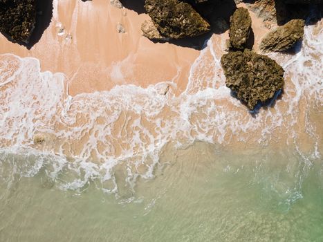Drone shot of a sandy beach with waves crashing on rocks, capturing nature's beauty.