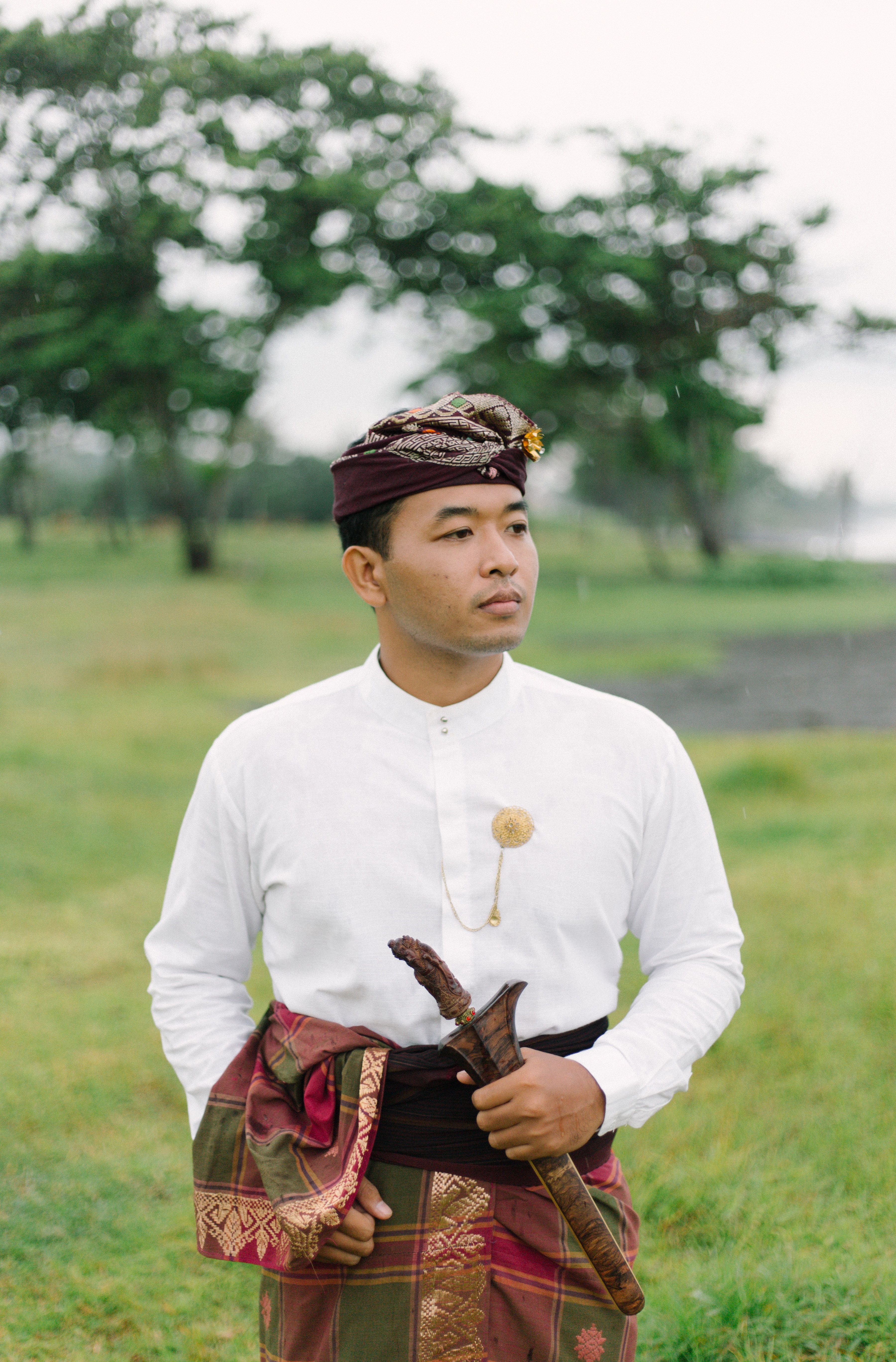 A man wearing traditional Balinese clothing stands in a lush green landscape, exuding cultural elegance.