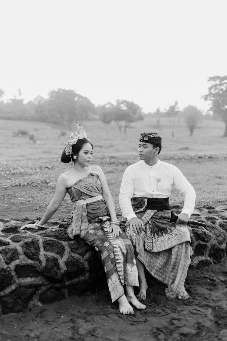 Black And White Photograph Of An Asian Couple Wearing Traditional Clothes Sitting On Clay Fence In An Orchard