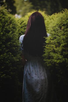 一位身着蓝色连衣裙的女子站在郁郁葱葱的灌木丛中，营造出神秘的户外场景