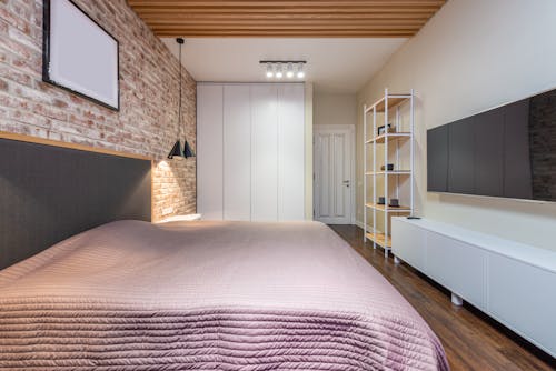 Bed with cover under empty panel on brick wall against television with black screen above cabinet at home