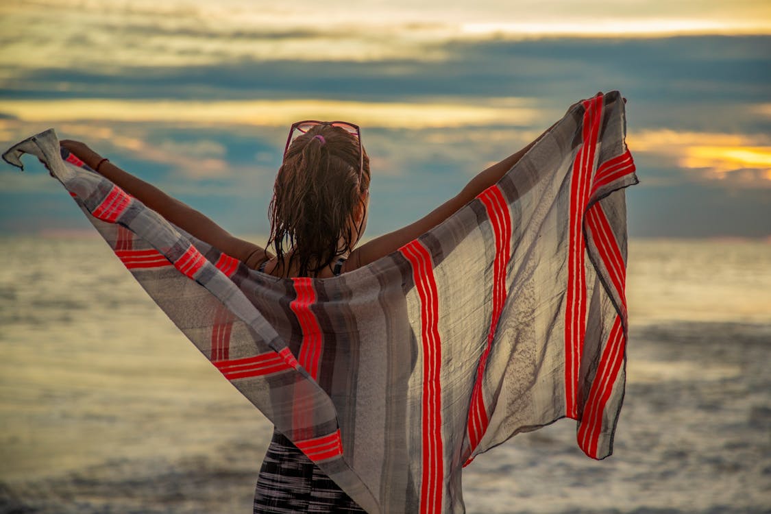 Woman Spreading Arms with Striped Red and Gray Scarf While Looking at the Sea at Sunset 
