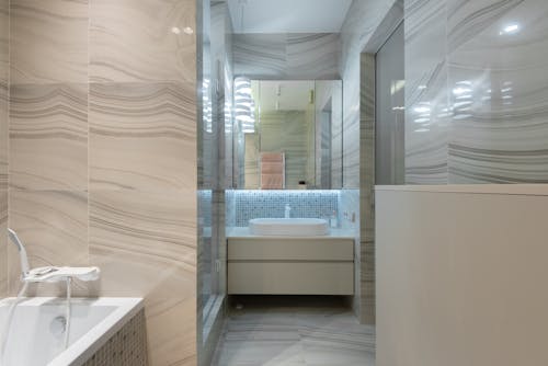Passage with tiled floor in contemporary bathroom with bath and washstand under mirror reflecting shiny lamps and towels at home