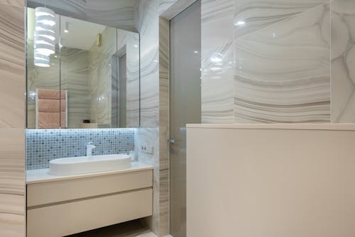 Modern bathroom interior with door against washstand between beige cabinet and mirror reflecting shiny lamps in house