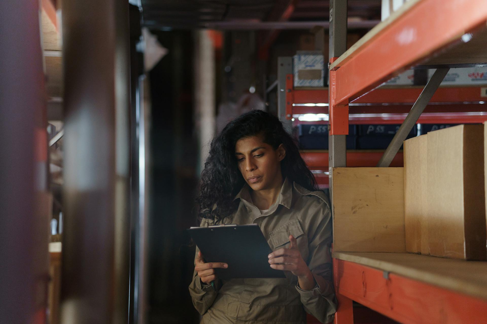 Woman in warehouse taking inventory with clipboard, focused and pensive.