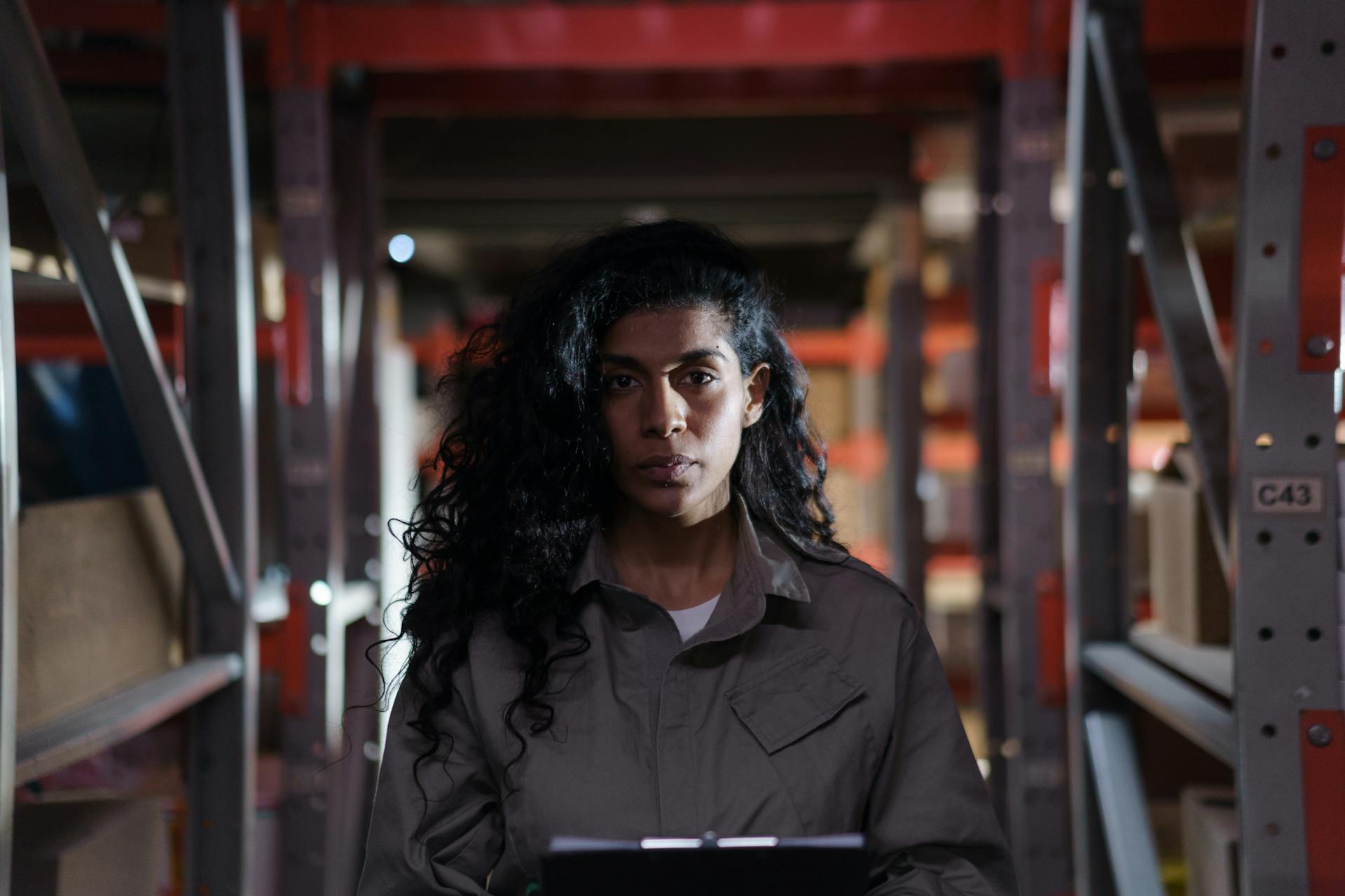 A woman holding a clipboard in a dimly lit warehouse aisle, focused on inventory management.