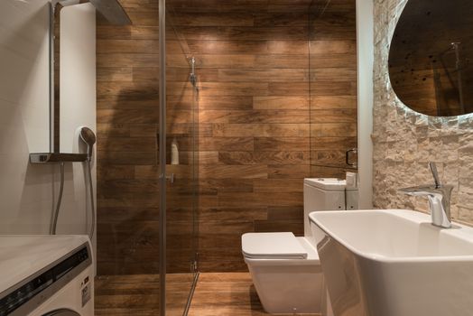 Stylish modern bathroom interior design with glass shower enclosure toilet bowl and sink under oval mirror