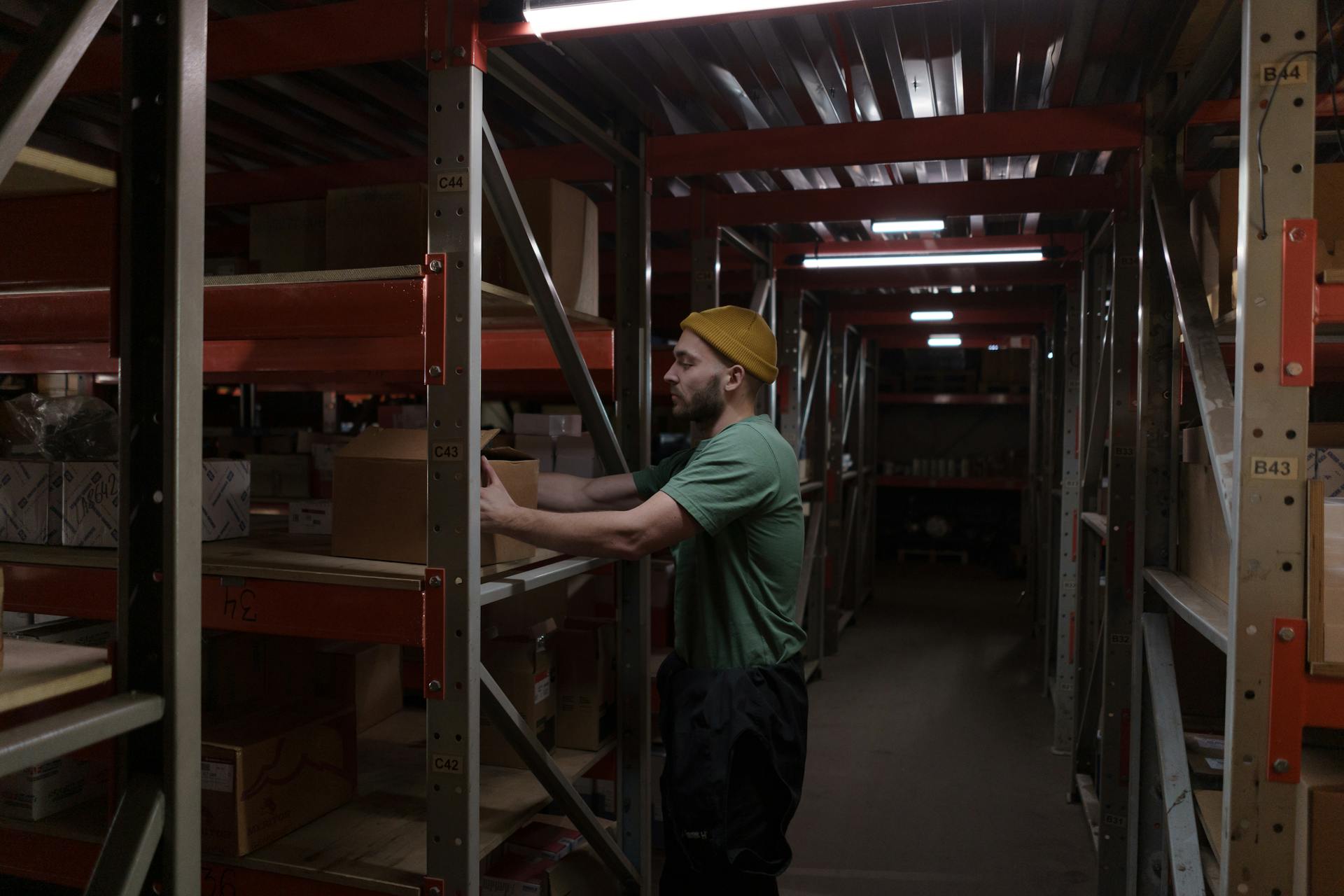 A man in a beanie and green shirt arranging boxes on shelves in a dimly lit warehouse storeroom.