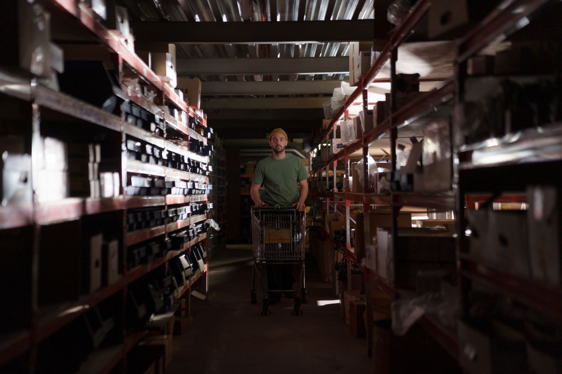 A man wearing a beanie pushes a cart through a dimly lit warehouse aisle.