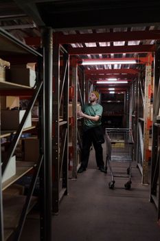 A man with a beanie and beard arranging items on a warehouse shelf with a cart nearby.