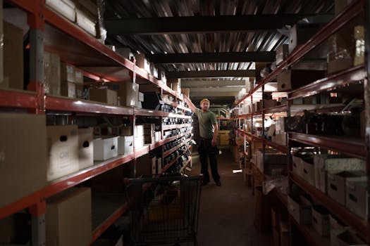 A worker organizing inventory in a dimly lit warehouse aisle full of shelves and boxes.