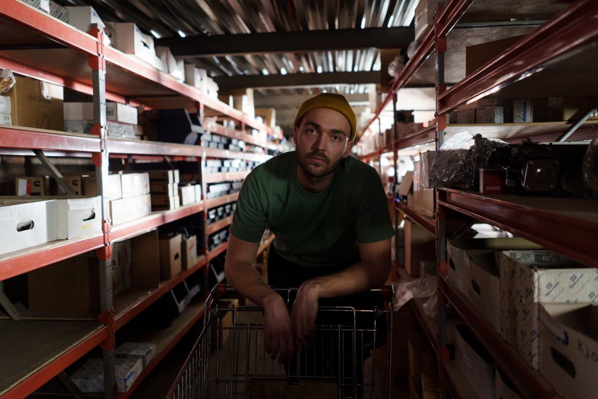 A worker leaning on a cart in a narrow parts storeroom aisle