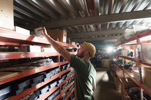 A man in a green shirt and yellow beanie organizing boxes in a warehouse aisle.