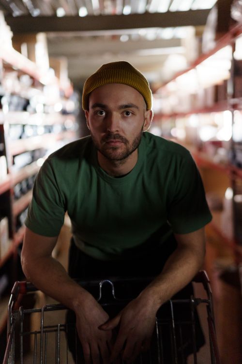 Free A thoughtful man wearing a green shirt and yellow beanie in a warehouse aisle. Stock Photo